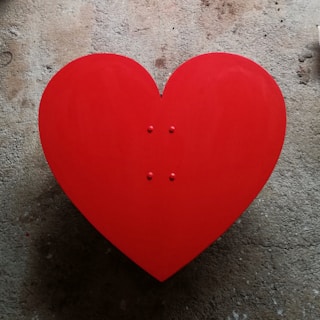 A large red heart shape placed on a textured concrete background. The heart is symmetrical and vibrant, standing out against the muted and rough surface.