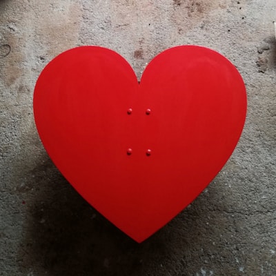 A large red heart shape placed on a textured concrete background. The heart is symmetrical and vibrant, standing out against the muted and rough surface.