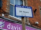 A blue and white sign reads 'Stand for 7 Taxis 24 Hours,' mounted on a rusty pole against a brick building. A window with closed blinds is visible in the background, and a shop sign with partial text 'davis...' in white on a purple background is beneath.