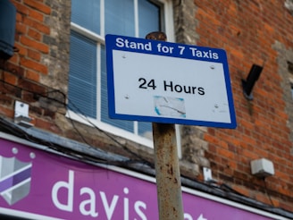 A blue and white sign reads 'Stand for 7 Taxis 24 Hours,' mounted on a rusty pole against a brick building. A window with closed blinds is visible in the background, and a shop sign with partial text 'davis...' in white on a purple background is beneath.