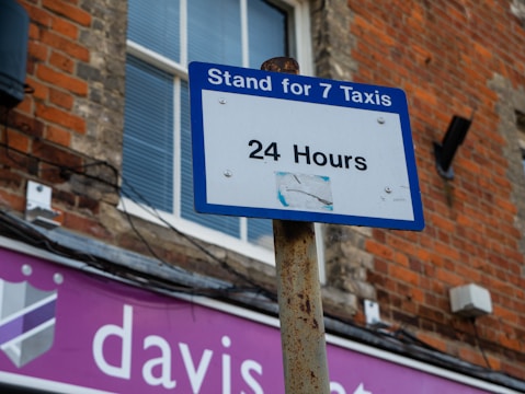 A blue and white sign reads 'Stand for 7 Taxis 24 Hours,' mounted on a rusty pole against a brick building. A window with closed blinds is visible in the background, and a shop sign with partial text 'davis...' in white on a purple background is beneath.
