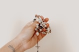 A hand holding a string of prayer beads, each composed of variously colored stones. A small cross hangs at the end of the string, and the person has a tattoo on their wrist.