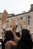 A group of people outdoors raising cardboard signs, one of which reads 'WHY LIVE IN FEAR'. The background shows a gray building with multiple windows. The individuals appear to be participating in a protest or demonstration.