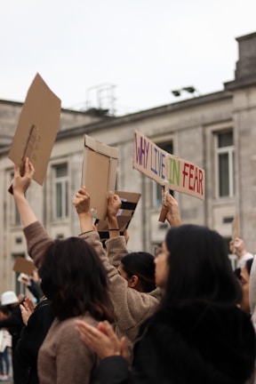 A group of people outdoors raising cardboard signs, one of which reads 'WHY LIVE IN FEAR'. The background shows a gray building with multiple windows. The individuals appear to be participating in a protest or demonstration.