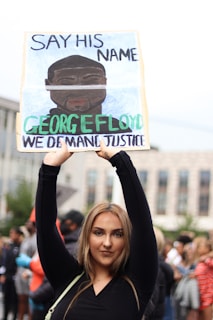 A person is holding up a protest sign with the text 'Say His Name George Floyd We Demand Justice' in a crowded outdoor setting. The background shows a blurred crowd of people, suggesting a protest or demonstration.