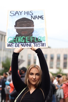 A person is holding up a protest sign with the text 'Say His Name George Floyd We Demand Justice' in a crowded outdoor setting. The background shows a blurred crowd of people, suggesting a protest or demonstration.
