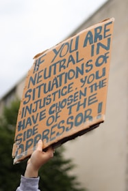 A hand is holding up a cardboard protest sign with bold, handwritten lettering. The sign has a message about neutrality and oppression, emphasizing a strong stance on social justice issues.