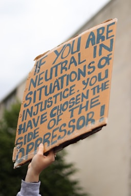 A hand is holding up a cardboard protest sign with bold, handwritten lettering. The sign has a message about neutrality and oppression, emphasizing a strong stance on social justice issues.