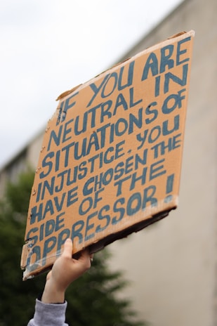 A hand is holding up a cardboard protest sign with bold, handwritten lettering. The sign has a message about neutrality and oppression, emphasizing a strong stance on social justice issues.