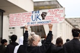 A group of people are participating in a protest, with one individual prominently holding a sign that reads 'The UK is not innocent' and lists several names. The setting appears to be an urban environment, with a focus on conveying a message of social justice.
