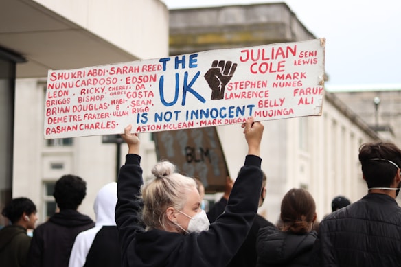 A group of people are participating in a protest, with one individual prominently holding a sign that reads 'The UK is not innocent' and lists several names. The setting appears to be an urban environment, with a focus on conveying a message of social justice.