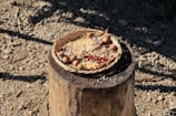 A woven basket sits on a wooden stump filled with various grains and seeds, including corn and small red beans. Surrounding the grains, small round objects resembling acorns are placed. The setting is outdoors on a dry, rocky surface with scattered stones and shadows cast across the ground.