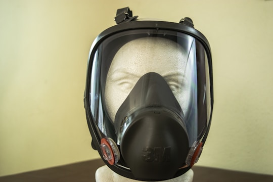 A mannequin head wearing a black full-face respirator mask is positioned against a plain wall. The mask features a clear protective visor and a filter on the front, designed for respiratory protection. The mannequin head appears to be made of a textured, white material.