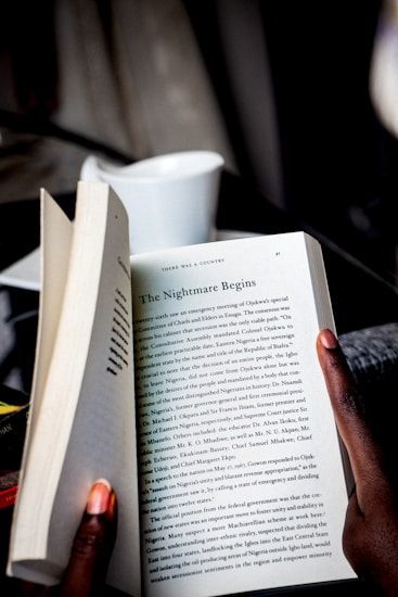 A person holds open a book, revealing a page with the title 'The Nightmare Begins.' The text discusses historical and political events. The person's fingers are visible, and a white cup is blurred in the background.