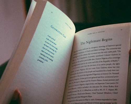 A person holding an open book, featuring a page from a text titled 'The Nightmare Begins' and another titled 'Generation Gap.' The lighting gives a soft, warm feel, highlighting the text and the hand holding the book.