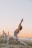 A woman is practicing yoga on a beach during sunset with a city skyline in the background. Her pose involves stretching her arms upward while one leg is bent forward and the other stretched back, creating a dynamic and balanced stance. The sky is a gradient of soft orange and blue, complementing the calm sea beside her.