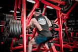 A person wearing a sleeveless shirt and cap works out on a piece of gym equipment. The setting contains numerous weights, with red-colored gym machines surrounding the individual. The environment is dimly lit with a focus on the exercise area.