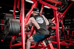 A person wearing a sleeveless shirt and cap works out on a piece of gym equipment. The setting contains numerous weights, with red-colored gym machines surrounding the individual. The environment is dimly lit with a focus on the exercise area.