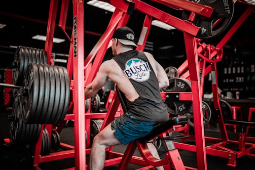 A person wearing a sleeveless shirt and cap works out on a piece of gym equipment. The setting contains numerous weights, with red-colored gym machines surrounding the individual. The environment is dimly lit with a focus on the exercise area.