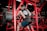 A person wearing a sleeveless shirt and cap works out on a piece of gym equipment. The setting contains numerous weights, with red-colored gym machines surrounding the individual. The environment is dimly lit with a focus on the exercise area.