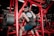A person wearing a sleeveless shirt and cap works out on a piece of gym equipment. The setting contains numerous weights, with red-colored gym machines surrounding the individual. The environment is dimly lit with a focus on the exercise area.