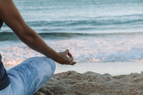 A person is seated in a meditative pose on a sandy beach, with the focus on their hand resting on their knee as they face the ocean. The gentle waves are visible in the background, evoking a sense of calm and tranquility.