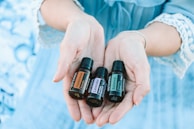 A pair of hands gently holds three small bottles of essential oils labeled 'Frankincense,' 'Lavender,' and 'Balance.' The background is a soft blue, matching the lace-trimmed sleeves of the person's clothing.