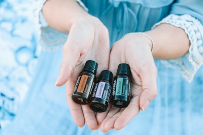 A pair of hands gently holds three small bottles of essential oils labeled 'Frankincense,' 'Lavender,' and 'Balance.' The background is a soft blue, matching the lace-trimmed sleeves of the person's clothing.
