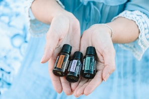 A pair of hands gently holds three small bottles of essential oils labeled 'Frankincense,' 'Lavender,' and 'Balance.' The background is a soft blue, matching the lace-trimmed sleeves of the person's clothing.