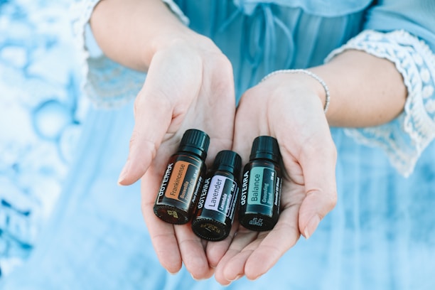 A pair of hands gently holds three small bottles of essential oils labeled 'Frankincense,' 'Lavender,' and 'Balance.' The background is a soft blue, matching the lace-trimmed sleeves of the person's clothing.