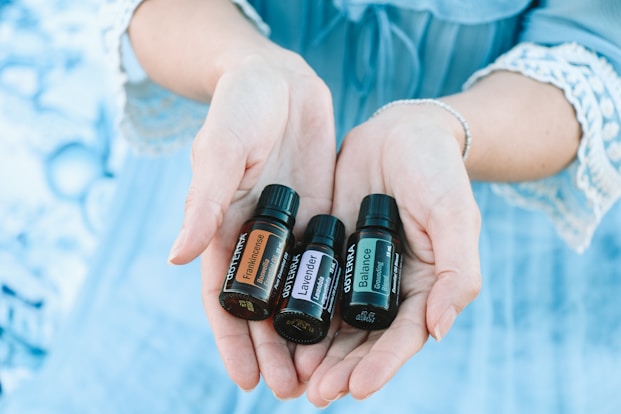A pair of hands gently holds three small bottles of essential oils labeled 'Frankincense,' 'Lavender,' and 'Balance.' The background is a soft blue, matching the lace-trimmed sleeves of the person's clothing.