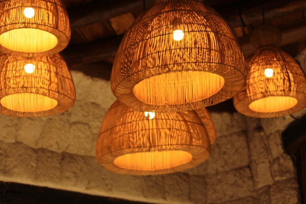 A series of woven lampshades hang from the ceiling, casting a warm and inviting glow. The lampshades are woven with natural fibers, creating intricate patterns that distribute light softly. The background consists of a textured stone wall, adding to the rustic and cozy atmosphere.