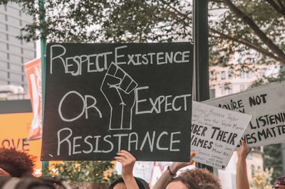 Multiple protest signs are being held up by people. The main sign in the center reads 'Respect Existence or Expect Resistance' with a fist graphic in the middle. Other signs reference names and call for attention to social issues. The background shows trees and some buildings with a somber and determined atmosphere.