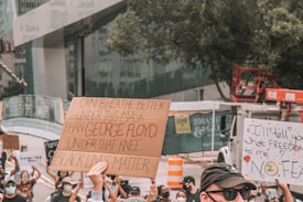 A group of people participating in a protest, some holding signs with messages advocating for justice and equality. One prominent sign reads 'I can breathe better under this mask than George Floyd under that knee Black Lives Matter'. People are wearing masks, suggesting a context during a period of concern for health and safety. Surrounding the protestors is an urban environment with trees and construction equipment visible.
