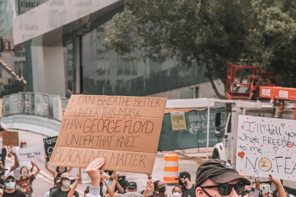 A group of people participating in a protest, some holding signs with messages advocating for justice and equality. One prominent sign reads 'I can breathe better under this mask than George Floyd under that knee Black Lives Matter'. People are wearing masks, suggesting a context during a period of concern for health and safety. Surrounding the protestors is an urban environment with trees and construction equipment visible.