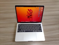 A laptop with a sleek silver finish is open on a wooden surface. Its screen displays a vibrant orange and red abstract floral wallpaper, and several application icons are visible on the desktop. The keyboard features a black background with white letters, and the laptop appears to be a compact model.