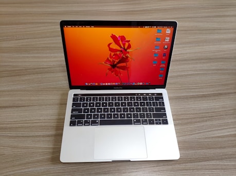 A laptop with a sleek silver finish is open on a wooden surface. Its screen displays a vibrant orange and red abstract floral wallpaper, and several application icons are visible on the desktop. The keyboard features a black background with white letters, and the laptop appears to be a compact model.