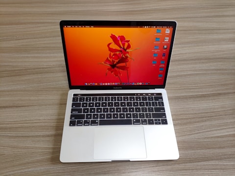 A laptop with a sleek silver finish is open on a wooden surface. Its screen displays a vibrant orange and red abstract floral wallpaper, and several application icons are visible on the desktop. The keyboard features a black background with white letters, and the laptop appears to be a compact model.