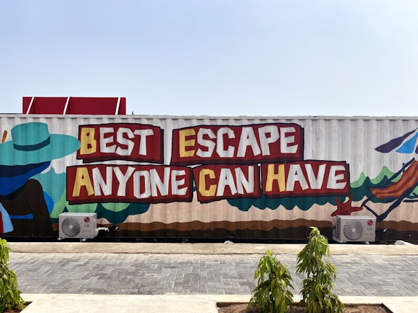 A large mural painted on the side of a shipping container with the words 'Best Escape Anyone Can Have' in bold red and yellow letters. The mural includes colorful depictions of a person wearing a blue hat and sitting in a deckchair, symbolizing relaxation or vacation. The container is situated outdoors, with small plants and paved ground in the foreground.