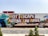 A large mural painted on the side of a shipping container with the words 'Best Escape Anyone Can Have' in bold red and yellow letters. The mural includes colorful depictions of a person wearing a blue hat and sitting in a deckchair, symbolizing relaxation or vacation. The container is situated outdoors, with small plants and paved ground in the foreground.