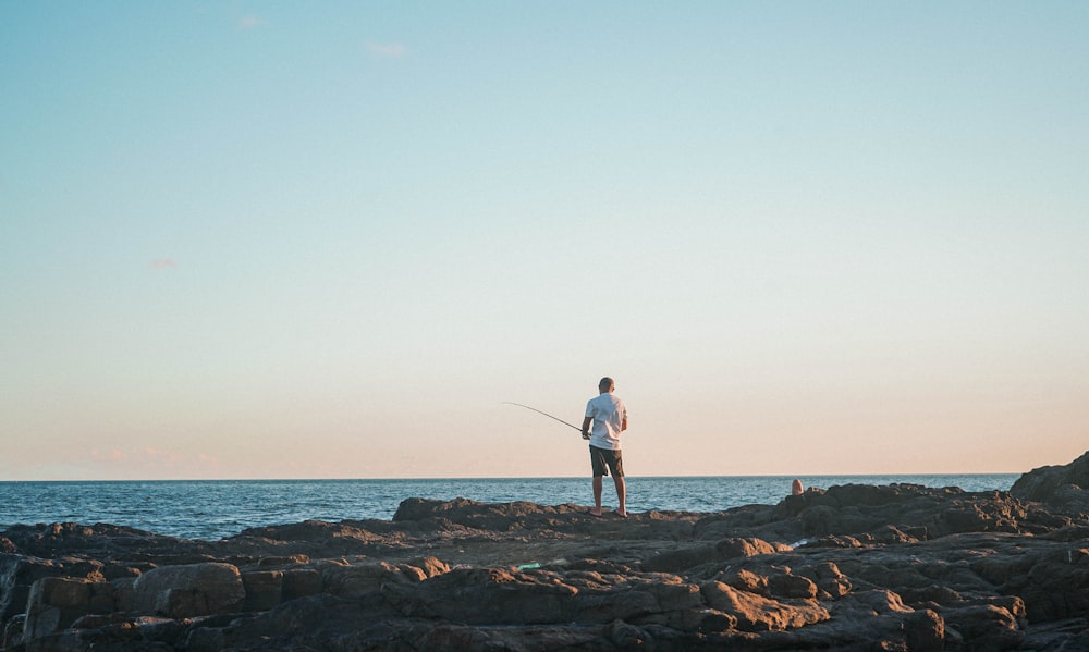 man in white shirt and blue denim jeans standing on rock near sea during daytime