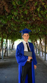 man in blue academic dress and academic hat standing near green tree during daytime