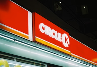 A brightly lit Circle K convenience store sign featuring a red and white color scheme, situated against a dark background. The sign includes distinctive branding and is illuminated, standing out prominently in the night setting.