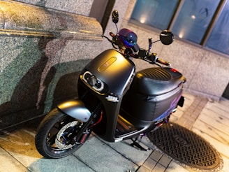 A sleek and modern black scooter is parked on a paved sidewalk, leaning against a stone wall. The scooter features a stylish design with a prominent headlight and well-crafted seat. The area is dimly lit, creating a subtle contrast and highlighting the scooter's glossy finish.