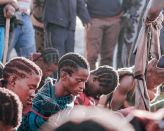 Several people with closely cropped hair are sitting and bowing their heads. They appear to be in some kind of communal or solemn gathering. Some individuals are wrapped in colorful, patterned blankets. Others are seen standing in the background, partially visible.