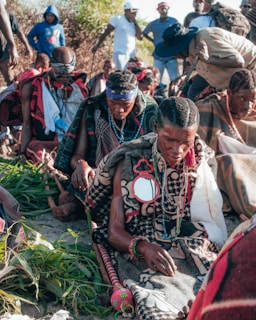 A group of people are gathered outdoors, seated on the ground, wearing traditional clothing and adorned with beads, necklaces, and headbands. They appear to be engaged in a ceremonial or cultural activity. In the background, more individuals are standing, some in casual clothing.