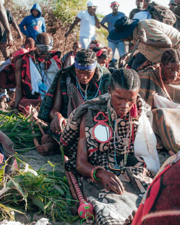 A group of people are gathered outdoors, seated on the ground, wearing traditional clothing and adorned with beads, necklaces, and headbands. They appear to be engaged in a ceremonial or cultural activity. In the background, more individuals are standing, some in casual clothing.