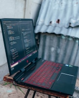 A gaming laptop with a red backlit keyboard is placed on a wooden stool. The screen displays a code editor filled with lines of colorful code. In the background, there is a corrugated metal sheet and a concrete wall.
