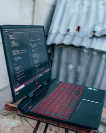 A gaming laptop with a red backlit keyboard is placed on a wooden stool. The screen displays a code editor filled with lines of colorful code. In the background, there is a corrugated metal sheet and a concrete wall.
