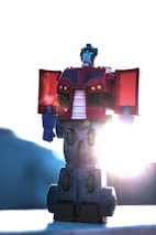 A toy robot figure stands against a bright background with the sun creating a glowing effect around its edges. The figure is predominantly red and blue, with mechanical features typical of a classic robot design. The sunlight creates a lens flare effect, enhancing the dramatic appearance of the toy.